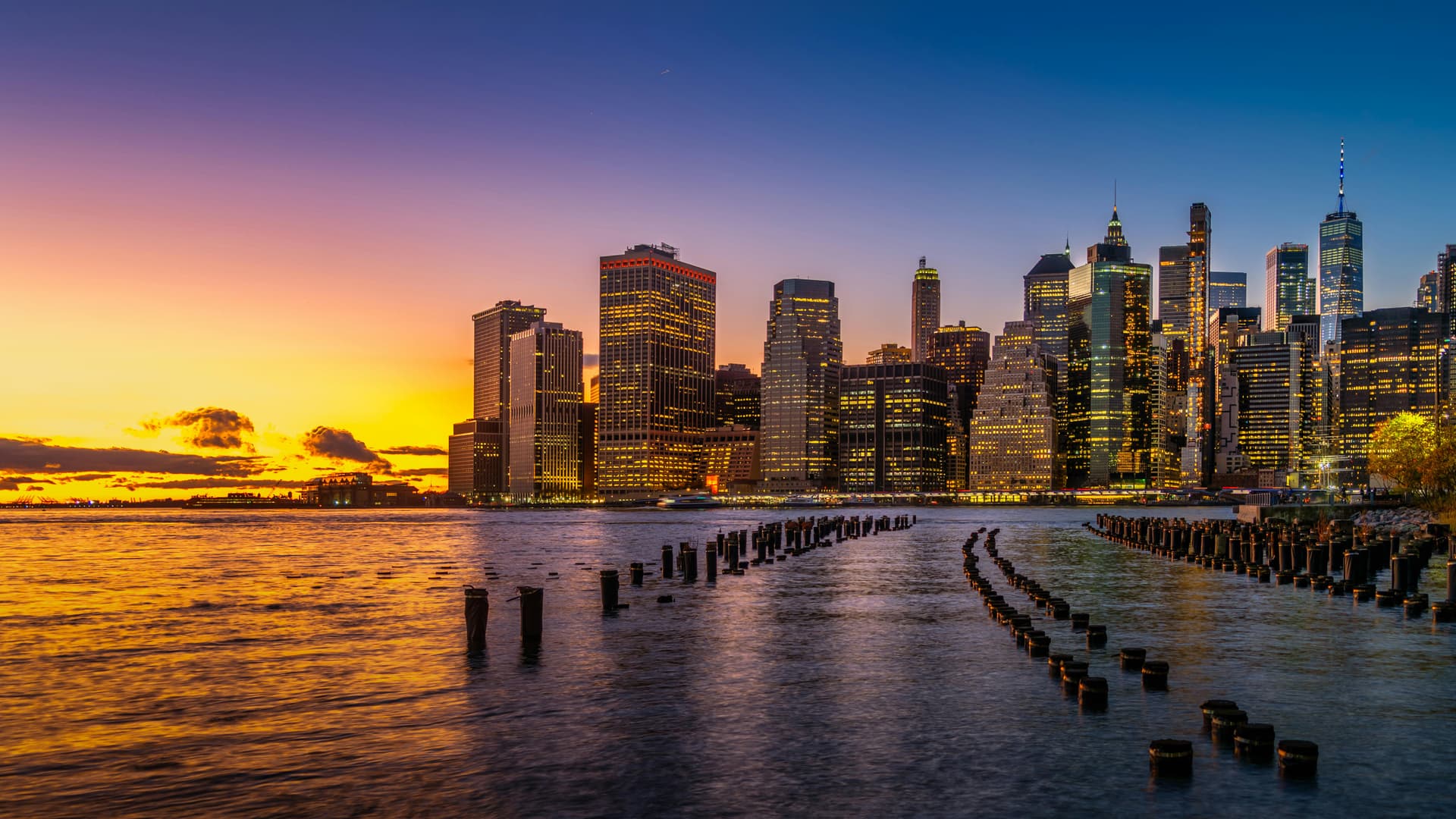 City financial-district skyline glowing at sunset across the water, old wooden pier pilings in the foreground