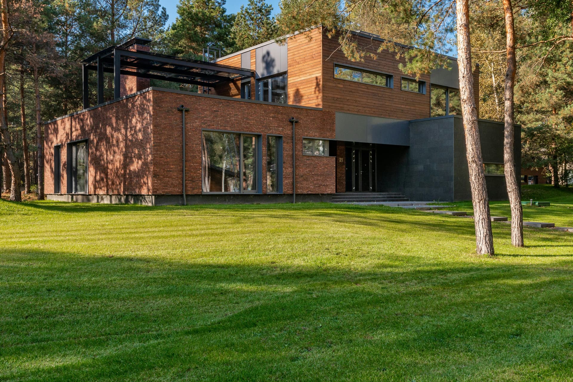 Modern brick-and-cedar contemporary home with floor-to-ceiling windows on a green lawn among tall pines