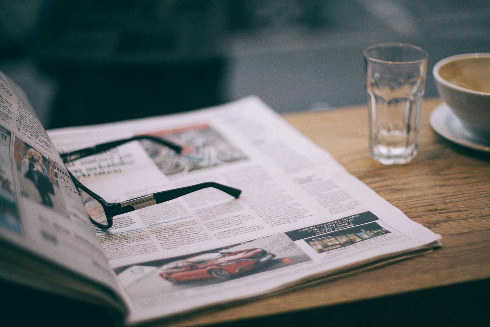 Folded newspaper with reading glasses, a glass of water, and a coffee cup on a wooden cafe table