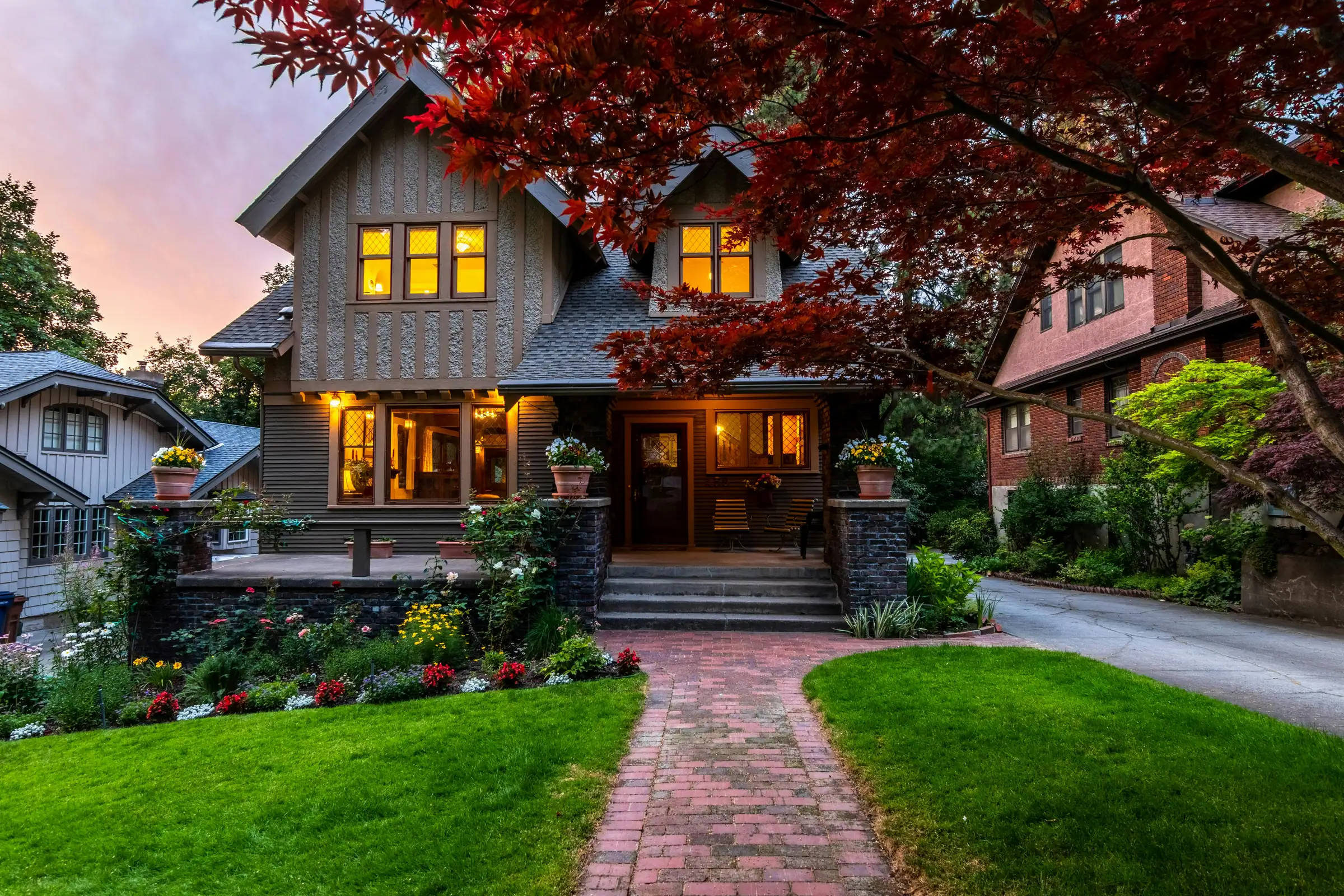 Craftsman-Tudor home glowing at dusk with timbered gables, flower gardens, and a brick walkway to the porch